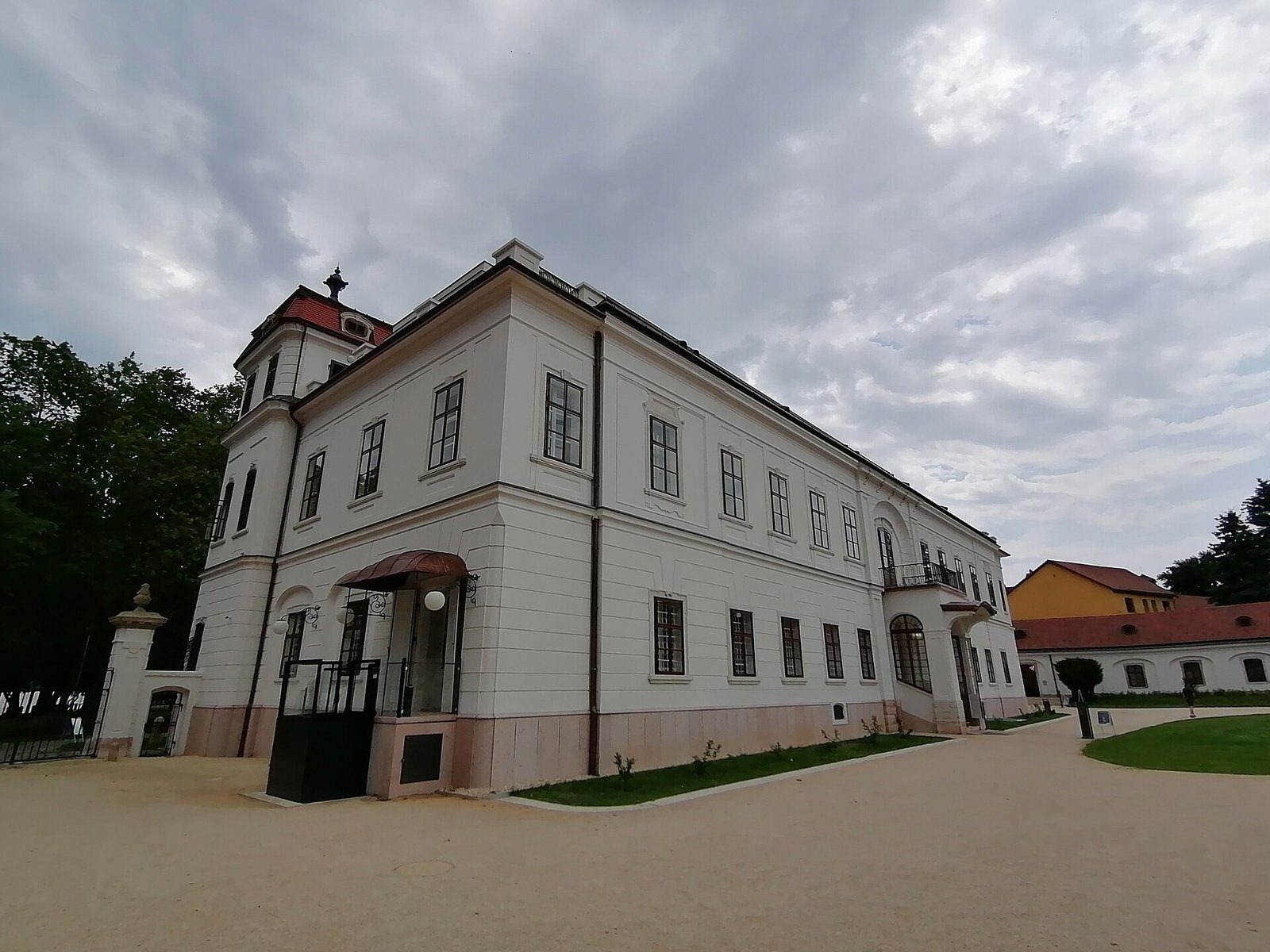 The restored Esterházy Palace in Tata, a long white two-story building with dark window frames and a small corner tower, on a gravel courtyard under a cloudy sky