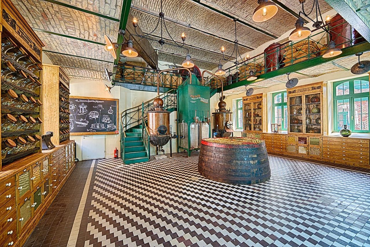 The old distillery hall at the Unicum House, with copper stills, a green column marked Zwack, a cask topped with dried herbs and wooden cabinets of herb jars under a brick-vaulted ceiling