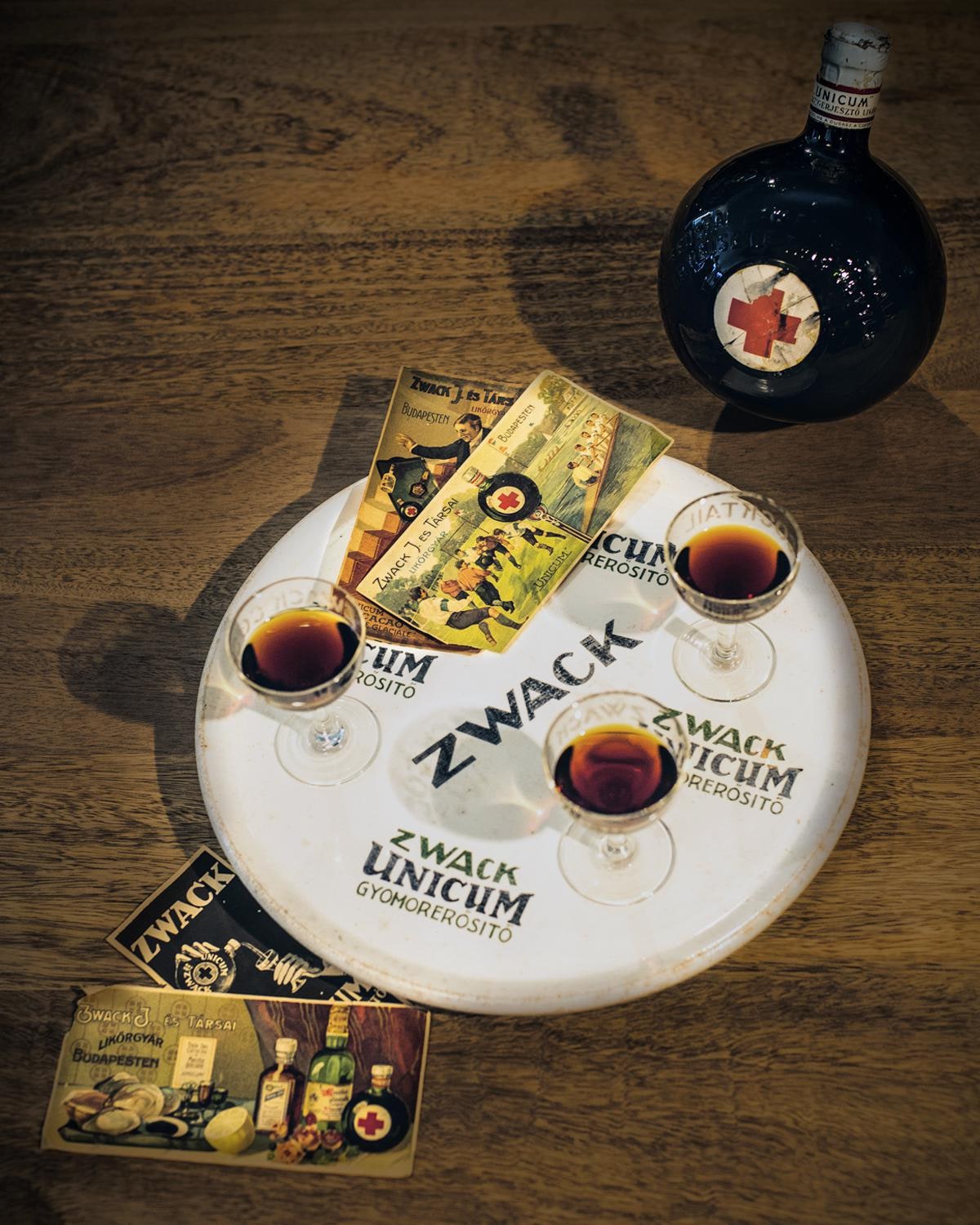 Three small glasses of Unicum on a white Zwack Unicum tray, beside a round Unicum bottle and vintage Zwack advertising cards