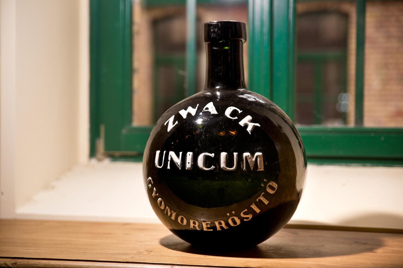An antique round dark glass bottle lettered Zwack Unicum Gyomorerősítő on a wooden windowsill in front of a green window frame