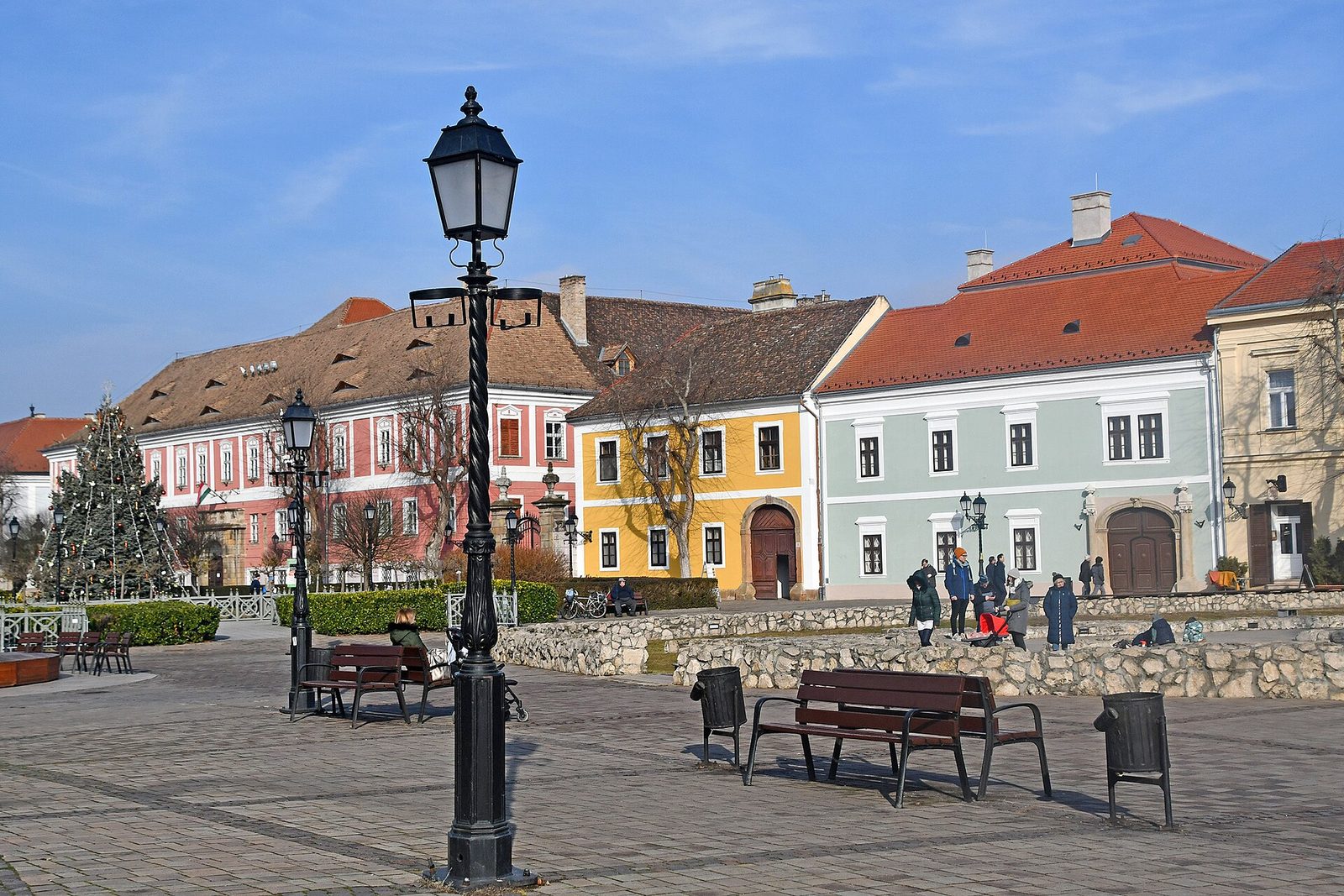 Március 15. tér w Vácu w słoneczny styczniowy dzień, pastelowe barokowe kamienice pod czerwonymi dachówkami, żeliwna latarnia, ławki i choinka