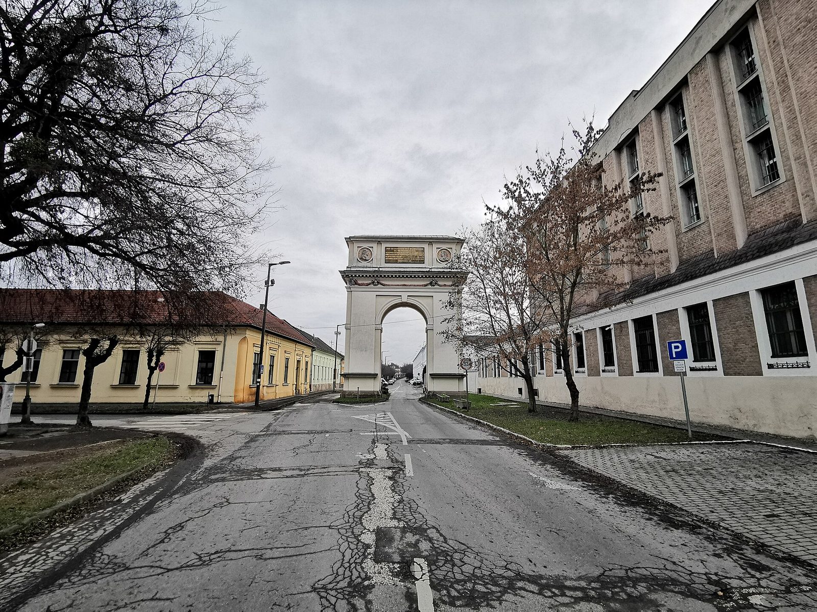 Kamienny łuk triumfalny w Vácu nad ulicą, widziany z daleka między bezlistnymi drzewami i niskimi budynkami w szary zimowy dzień