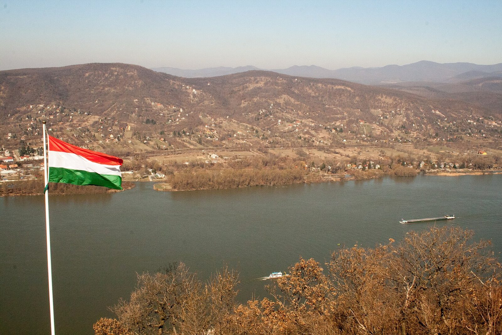 Dunaj widziany z Cytadeli w Visegrádzie, z węgierską flagą na pierwszym planie, barką na rzece i zalesionymi wzgórzami Nagymaros na drugim brzegu