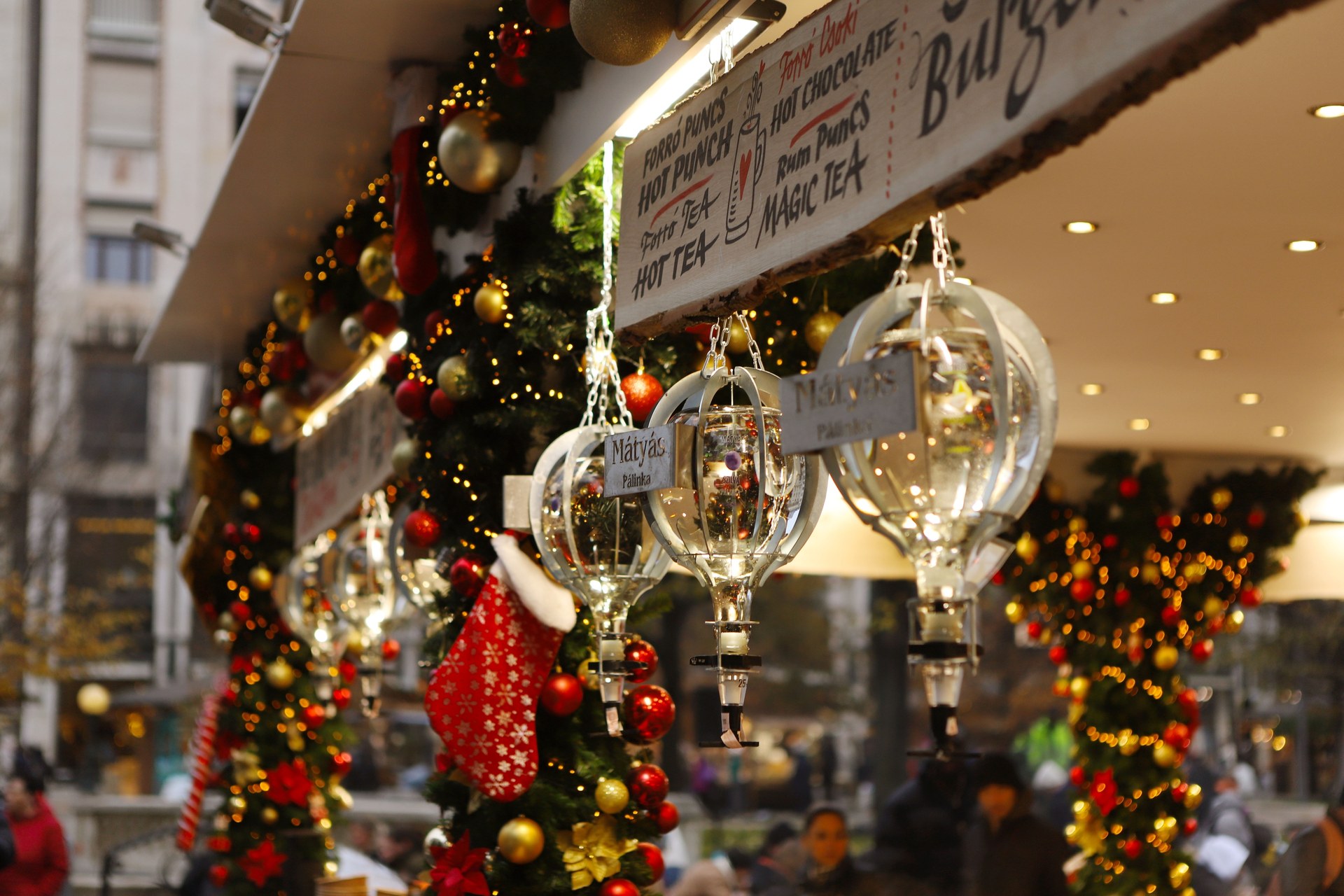 Glass pálinka dispensers with Mátyás Pálinka labels and a Christmas stocking hanging from a decorated stall selling hot drinks at the 2025 Vörösmarty tér Christmas market